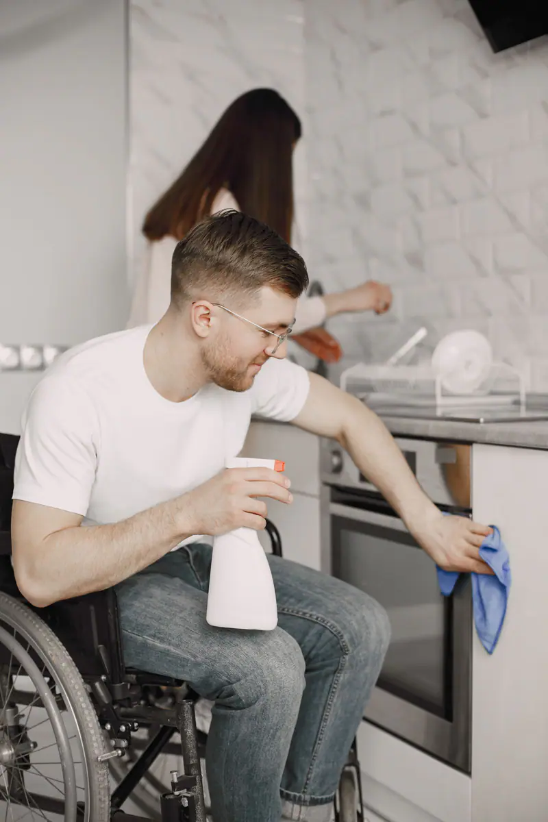 A wheelchair user cleaning a kitchen surface with spray and a cloth.