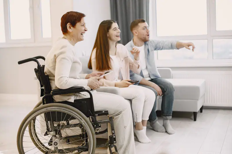 An older wheelchair user laughing at home with two smiling family members or support people.