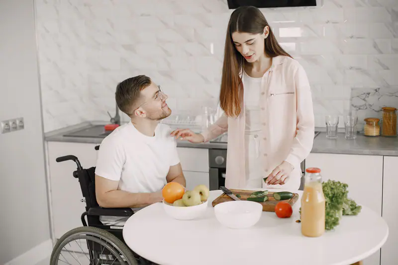 A wheelchair user learning food preparation with support in a home kitchen.