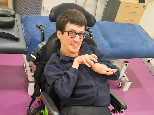 A smiling man with glasses seated in a green powered wheelchair.